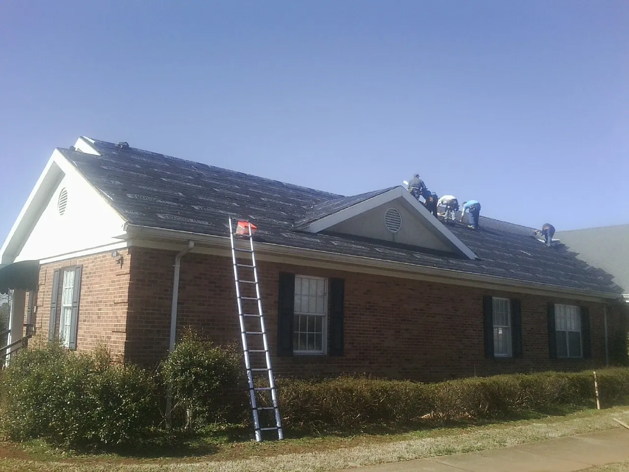 Crew tearing shingles off a brick ranch roof with a ladder set — Metal Roof Repair work by our crew in Bradley